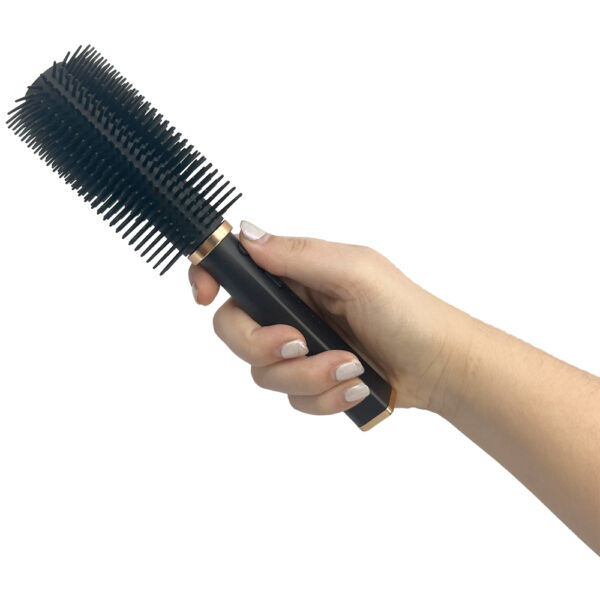 The image features a hand holding a black round hairbrush with dense bristles, designed for styling and blow-drying hair. The handle of the brush is sleek and black with a gold accent near the base of the bristles, indicating a touch of elegance in its design. The hand holding the brush has neatly manicured nails painted in a light,