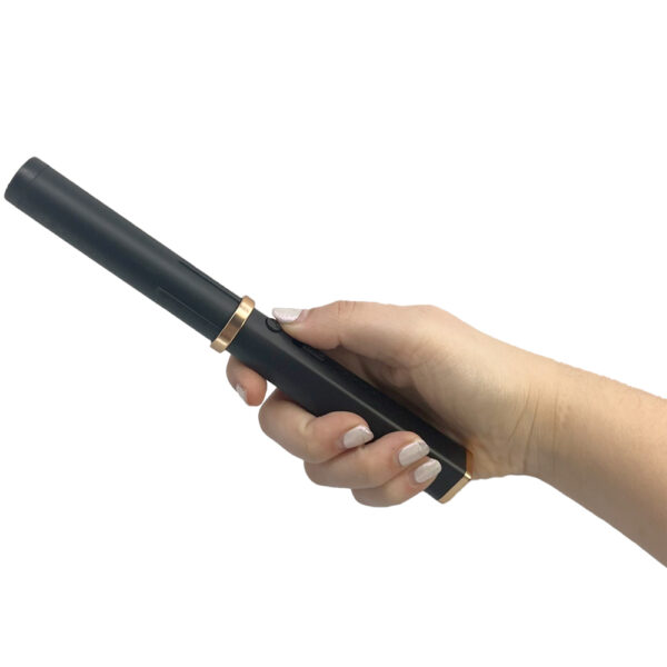 This image features a hand holding a black electronic device that resembles a sleek, cylindrical wand or pen. The device has a smooth matte black finish with a gold ring near its middle, adding a touch of elegance and modern design. The hand, with neatly manicured nails painted in a shimmering nude color, is positioned to grip the device comfortably. The background