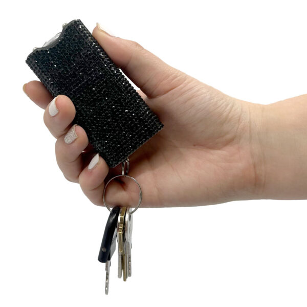 A person's hand is holding a small, black, rhinestone-covered self-defense stun gun keychain. The shiny surface of the device is studded with black crystals, giving it a glamorous appearance. It is attached to a keyring with several keys hanging, including a black plastic key fob and metallic keys. The hand has manicured nails, with