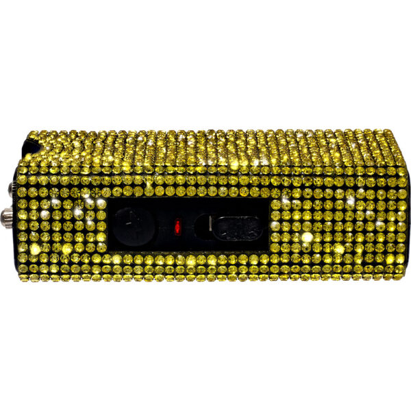 The image shows a rectangular taser adorned with bright yellow rhinestones, giving it a sparkling, eye-catching appearance. The surface is completely covered in these glittering embellishments, which are meticulously arranged in a uniform pattern. The device features a black panel on one side that houses two buttons and a switch, used for operating the taser. The contrast