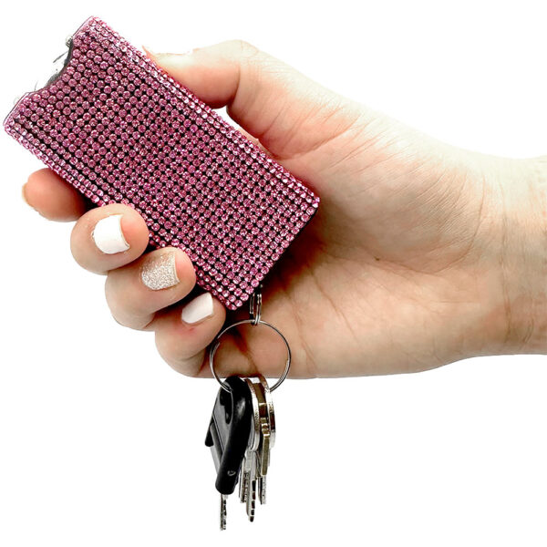 A hand is holding a stylish pink rhinestone-covered keychain in close-up. The keychain features a rectangular shape and is adorned with sparkling rhinestones that create a shimmering effect. Attached to the keychain are several keys, including a black plastic key and metal keys. The hand has neatly manicured nails painted in white, with one nail displaying a