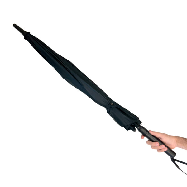 A black, compact umbrella is held by a hand against a white background. The umbrella is fully closed, showing its sleek, cylindrical shape. The handle is black and has a wrist strap attached, providing ease of carrying. The canopy fabric is neatly wrapped and secured with a snap button. This image highlights a portable and practical rain accessory, ideal for easy