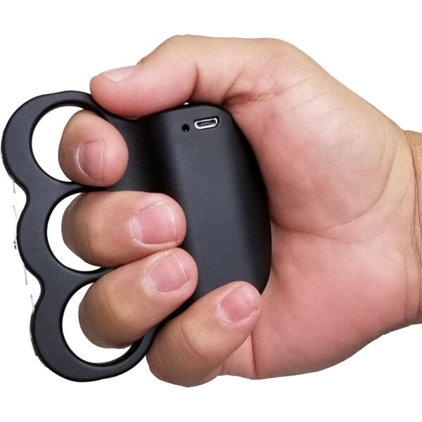 A close-up image depicts a hand gripping a black electronic knuckle-shaped device, likely a self-defense tool or stun gun. The device is snugly fitted around the person's fingers, with a smooth, matte finish. A small charging port is visible on its body, indicating rechargeable functionality. The image clearly showcases the ergonomic design, intended for easy handling and